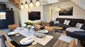 A modern living room with a dark sofa, a dining table set for four, and a large TV mounted on the wall.