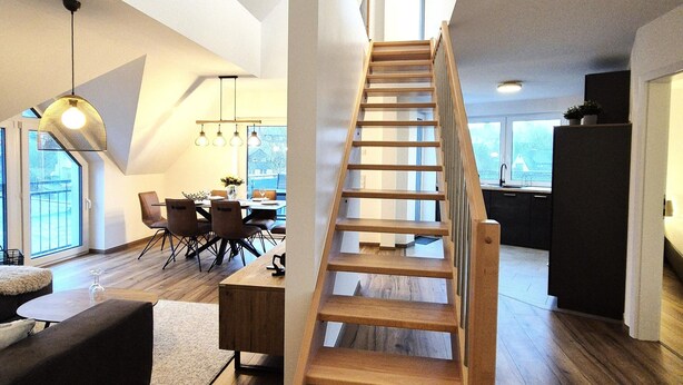 A modern interior with a wooden staircase, a dining area with chairs and a table, and a kitchen in the background.
