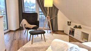 A modern bedroom with a large window, a chair with a throw blanket, a bedside table with books, and a rolled towel on the bed.