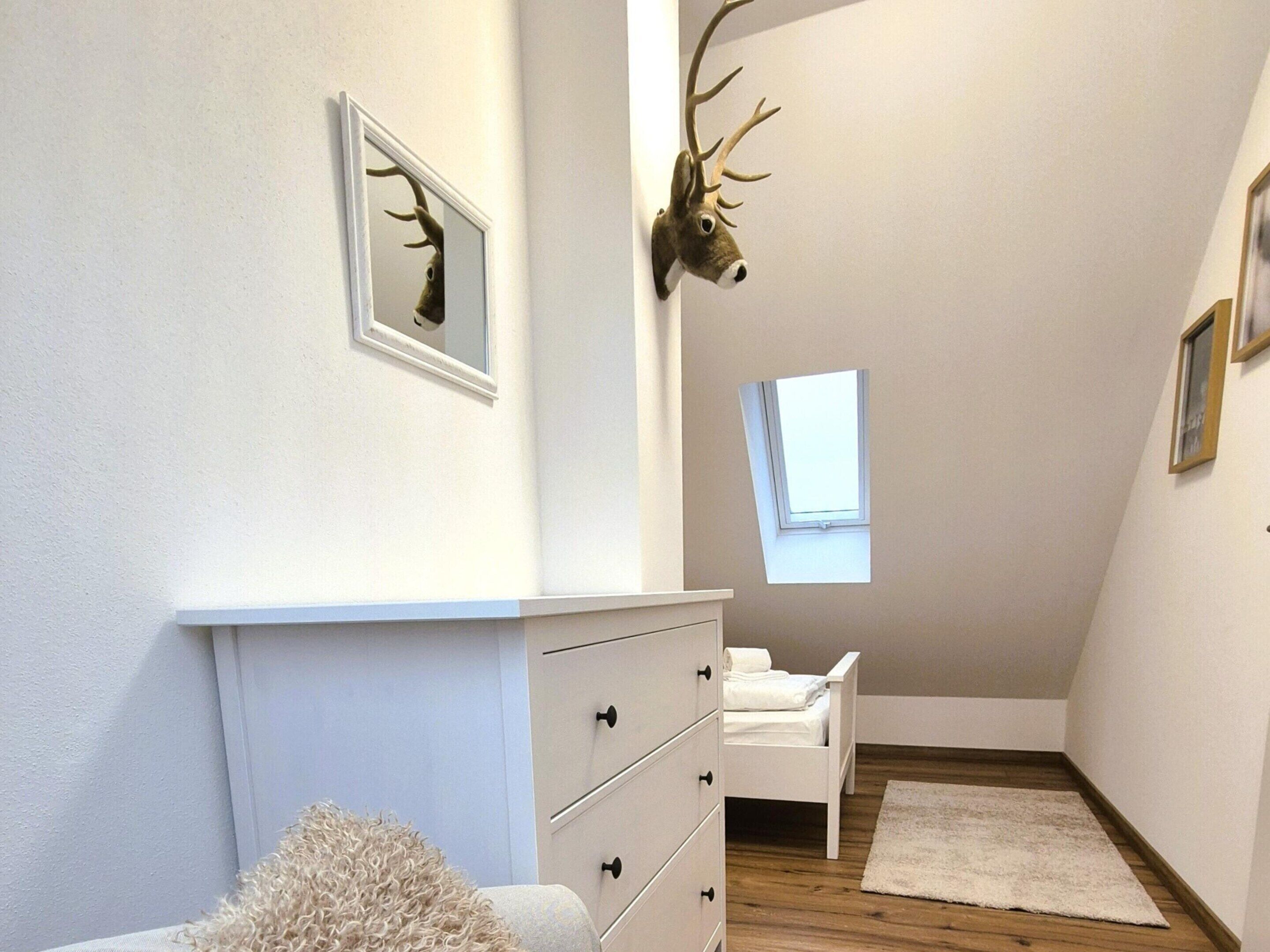 Een slaapkamer met een witte dressoir, een bed, een raam en een hertenhoofd aan de muur.