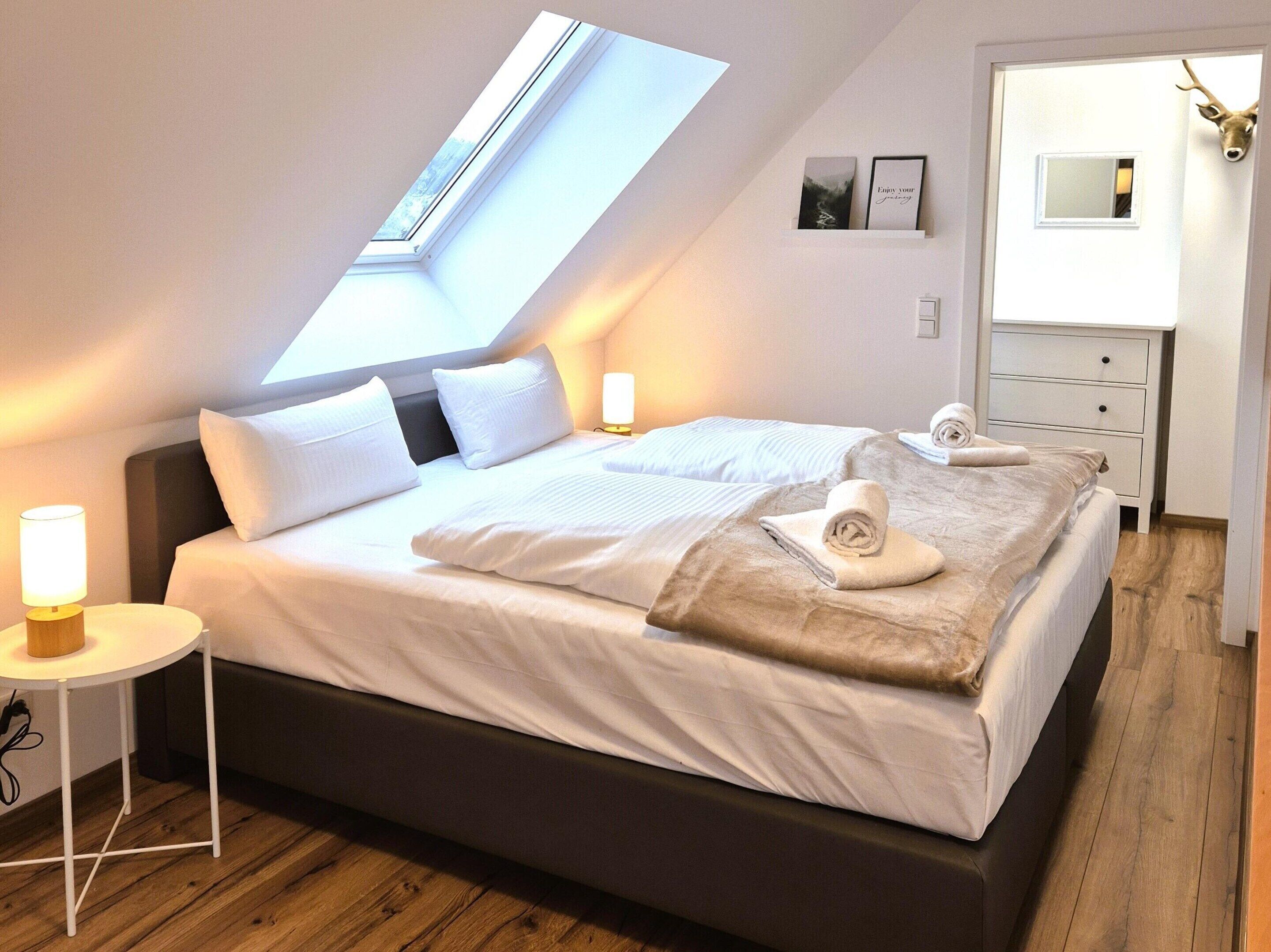 A modern bedroom with a skylight, a bed with white linens, a bedside table with a lamp, and a wooden floor.