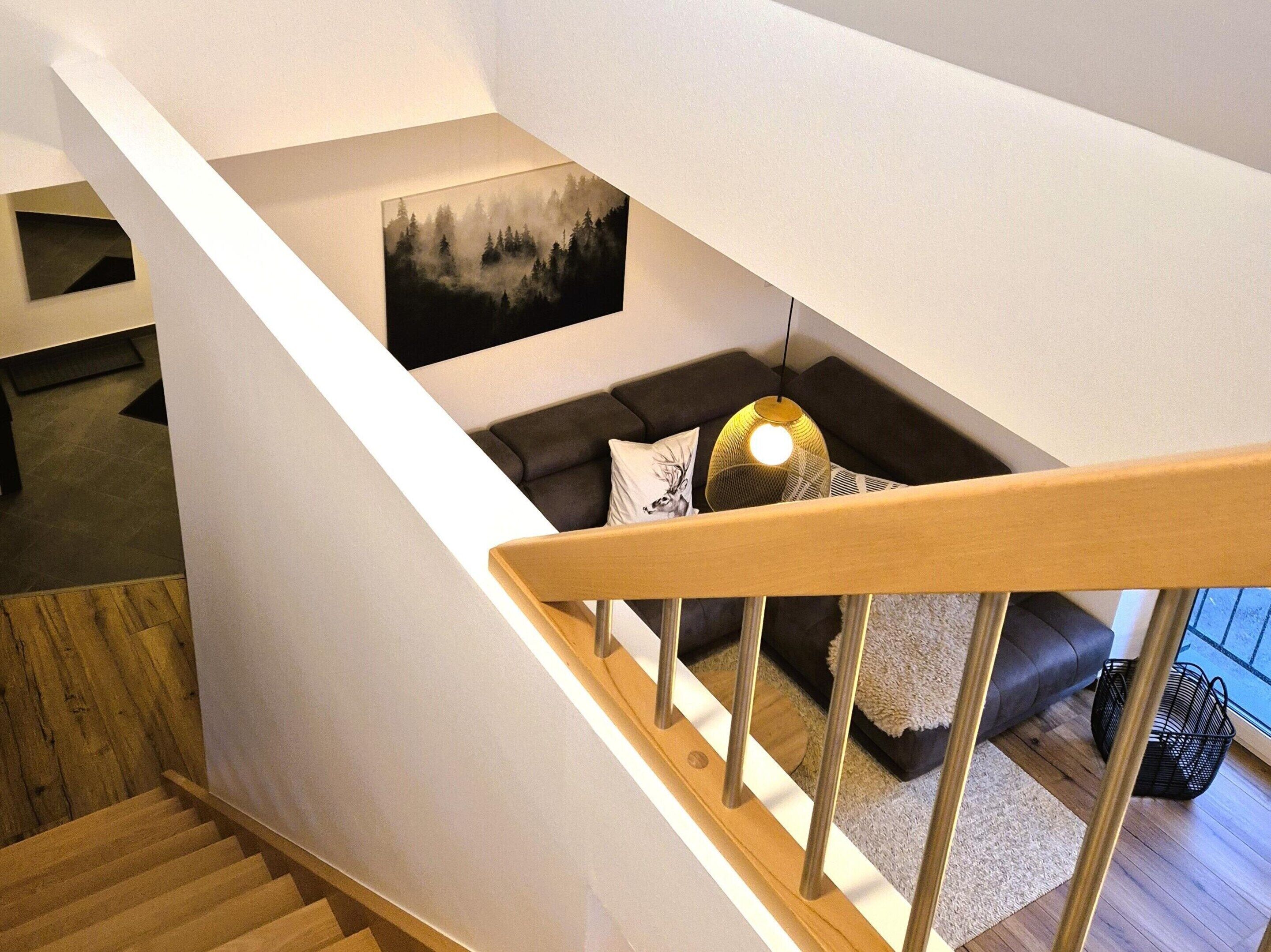 A modern staircase with wooden handrails leading to a living area with a dark sofa, a hanging light fixture, and a framed picture on the wall.
