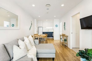 A modern living room with a grey sofa, a dining table with chairs, a kitchen in the background, and a TV mounted on the wall. - Modern 2BR in CBD w/ Full Amenities- Zodiak Stays (Auckland)