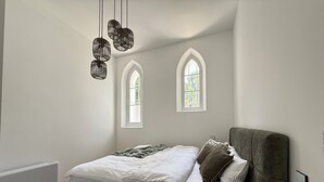 Un dormitorio con cama, dos ventanas, un radiador y una mesita de noche con un florero.