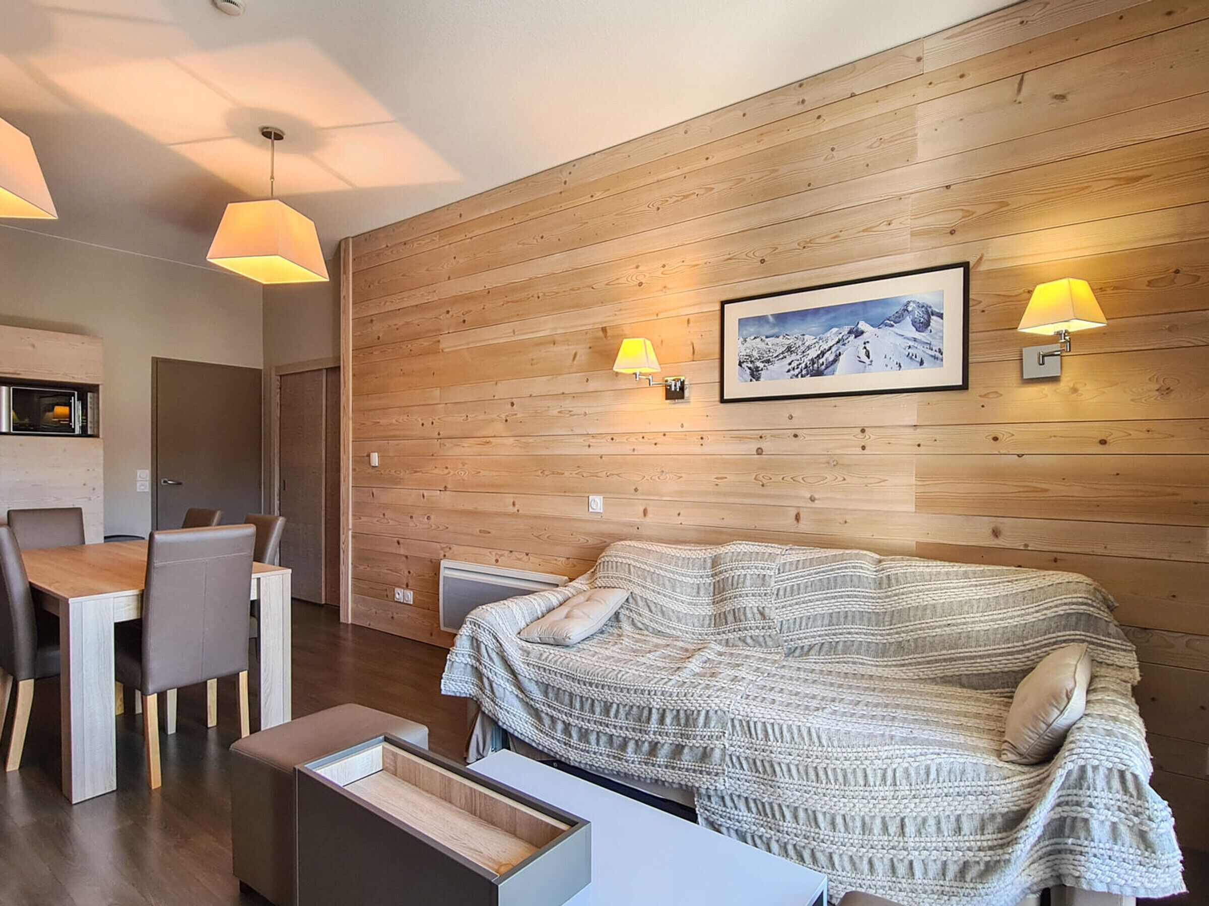 A modern room with a wooden wall, a bed with a knitted blanket, a dining table with chairs, and two wall-mounted lamps.