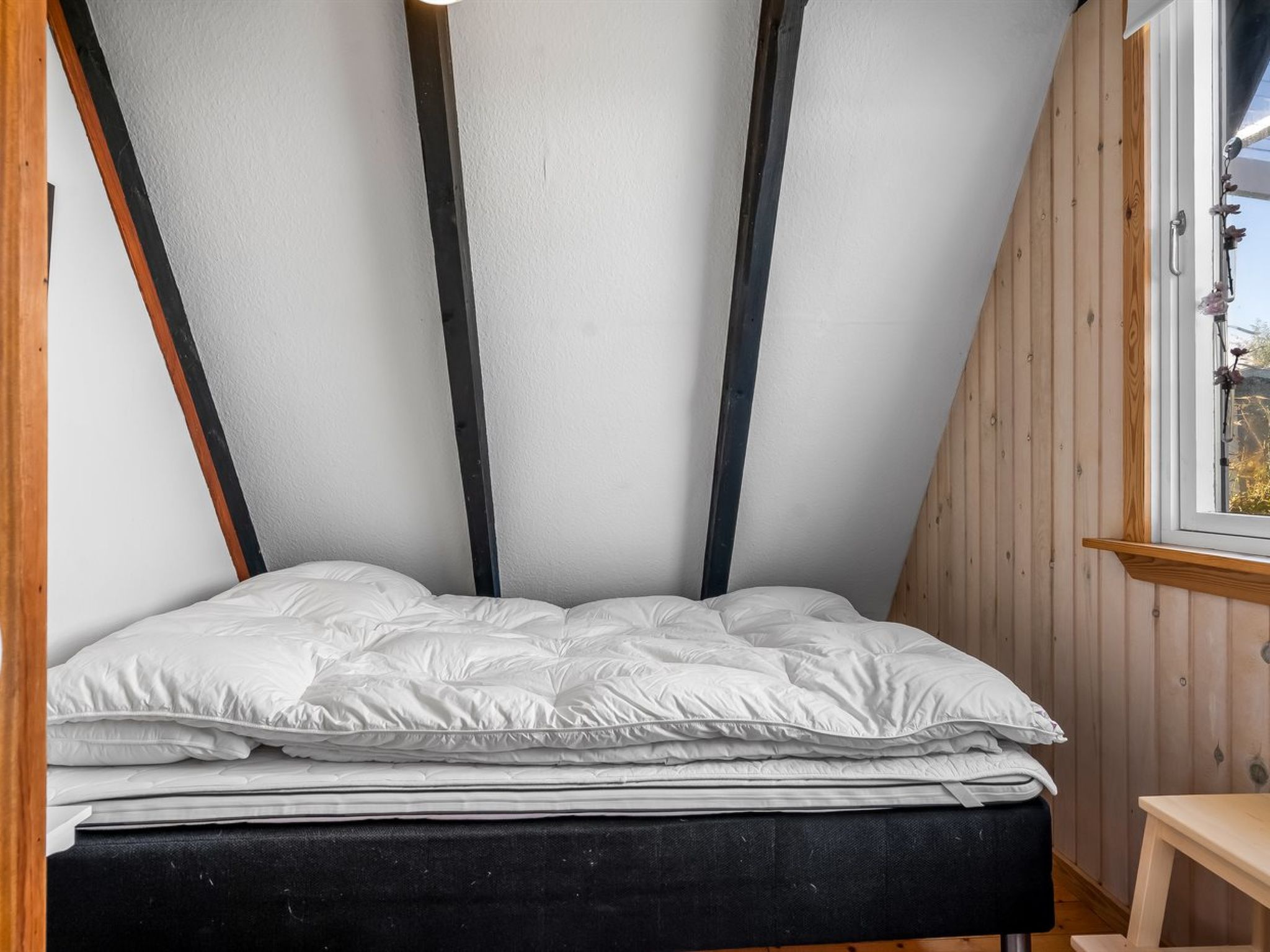 Habitació petita amb sostre inclinat, llit, parets de fusta i finestra.
