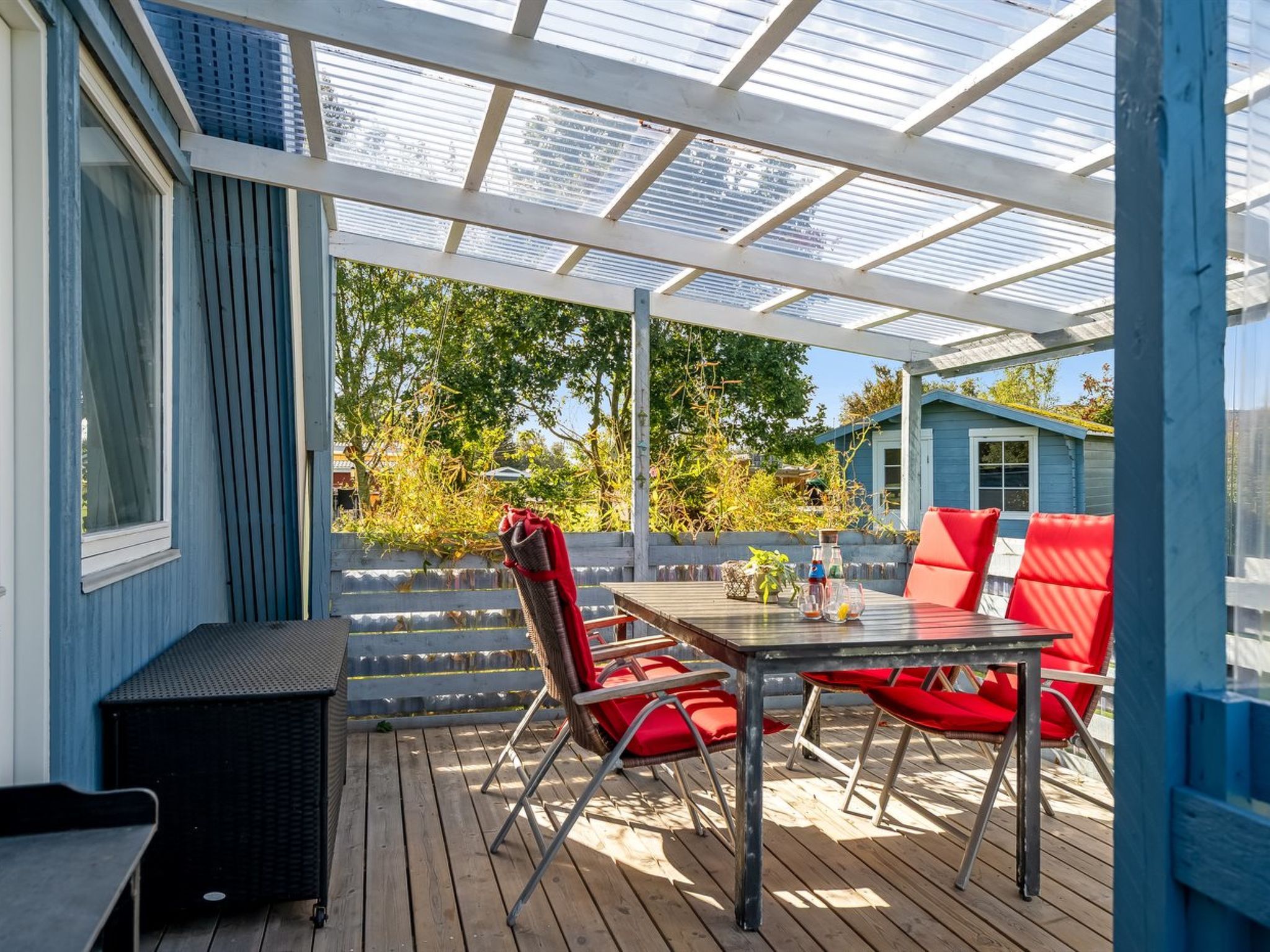A covered outdoor patio with a table and red chairs, a small black bench, and a wooden deck.
