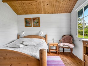 Ein Schlafzimmer mit einem hölzernen Bett, einem Sessel, einem Nachttisch und einem Fenster mit Blick auf Grünflächen.