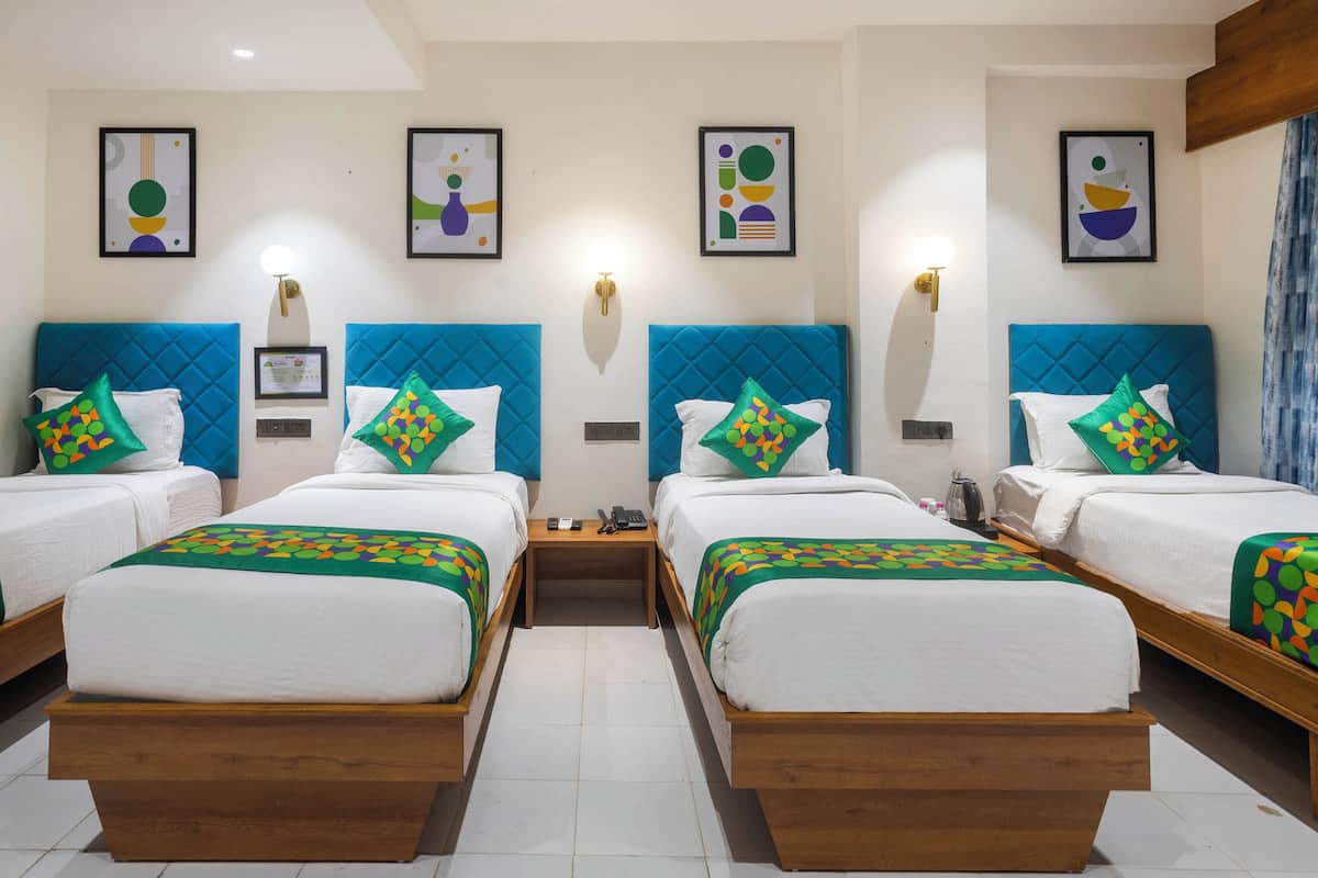 A hotel room with four beds, each with a blue headboard and colorful patterned bedspreads. There are wall-mounted light fixtures and framed artwork above each bed.