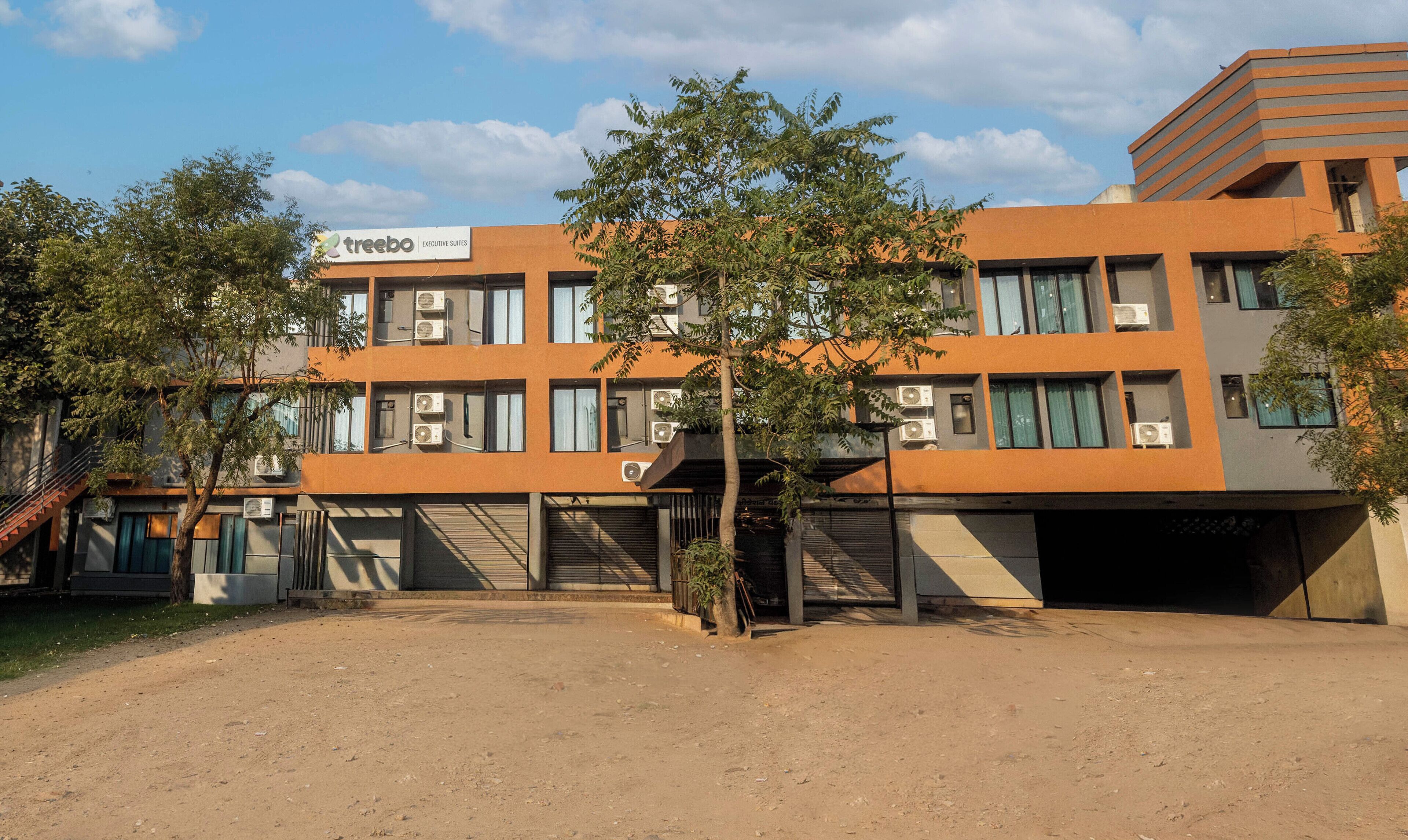Un edificio moderno con diverse unità di condizionamento, un cartello con la scritta "treebo Executive Suites" e un'area a terra sterrata con alberi.