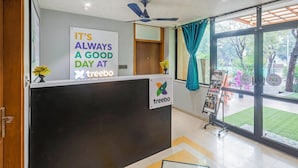 A reception area with a sign that reads "IT'S ALWAYS A GOOD DAY AT treebo" and a logo. There's a ceiling fan, a window with curtains, and a display rack with magazines. - Treebo Executive Suites (Gandhinagar)