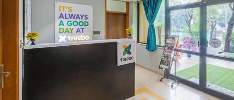 A reception area with a sign that reads "IT'S ALWAYS A GOOD DAY AT treebo" and a logo. There's a ceiling fan, a window with curtains, and a display rack with magazines.