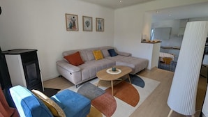 Un salon moderne avec un canapé gris, un fauteuil bleu, une petite table et un radiateur blanc. Trois œuvres d’art encadrées ornent le mur, et on aperçoit un coin cuisine au fond.