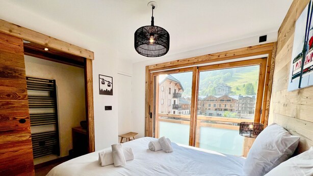 A bedroom with a large bed, a wooden door, a small table, and a view of buildings through a window.