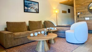 A cozy living room with a sofa, a round wooden coffee table, and a white armchair. There's a painting of mountains on the wall and a lamp on the floor.