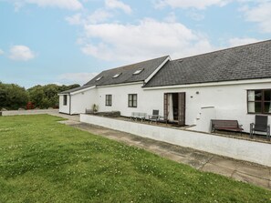 A white building with a grey roof, a paved driveway, and a grassy lawn with white flowers. - Florence Cottage (Holyhead)