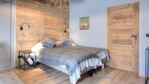 Un dormitorio con techo de madera, una cama con manta gris, dos almohadas, una mesita de noche, una lámpara de pared y una puerta.