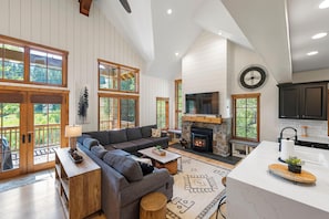 A spacious living room with a fireplace, a large sofa, and a kitchen area with dark cabinets and a white countertop.