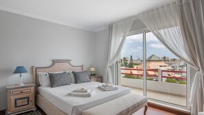 Un dormitorio con una cama grande, una mesita de noche, una lámpara, una ventana con cortinas y vistas a edificios.