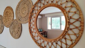 Una habitación con un espejo circular grande y un marco tejido, tres tapices tejidos más pequeños en la pared, y una ventana con vistas a follaje exterior.