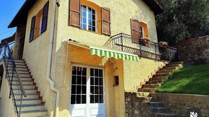 Una casa de dos pisos con una escalera de piedra que lleva a la entrada, un balcón con barandilla metálica y un pequeño porche.