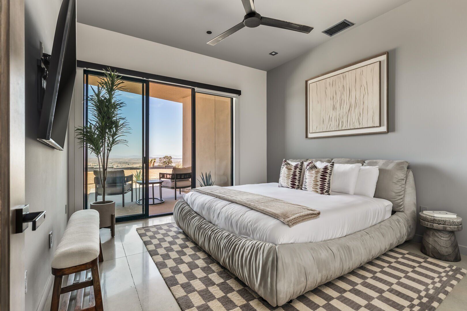 A modern bedroom with a large bed, a ceiling fan, a TV, and a balcony with a view of the landscape.
