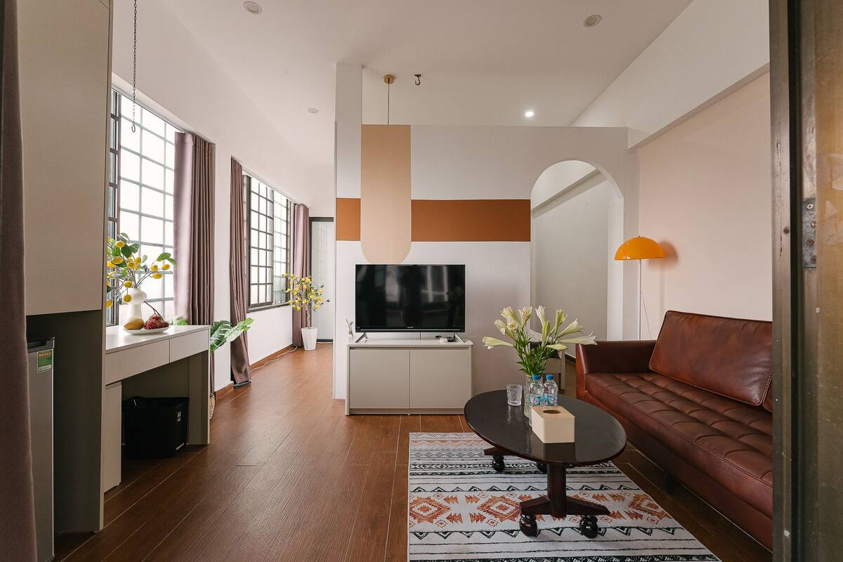 A modern living room with a brown leather sofa, a coffee table, and a flat-screen TV. There is a dining area with a white table and chairs, and a kitchen area with a refrigerator and cabinets.