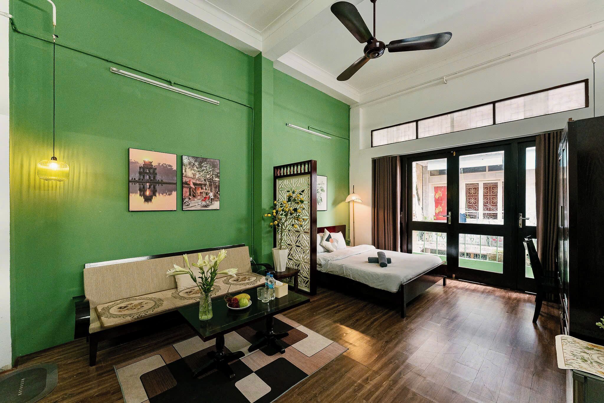 A hotel room with a green wall, a bed, a sofa, a desk with a vase of flowers, and a view through the window.