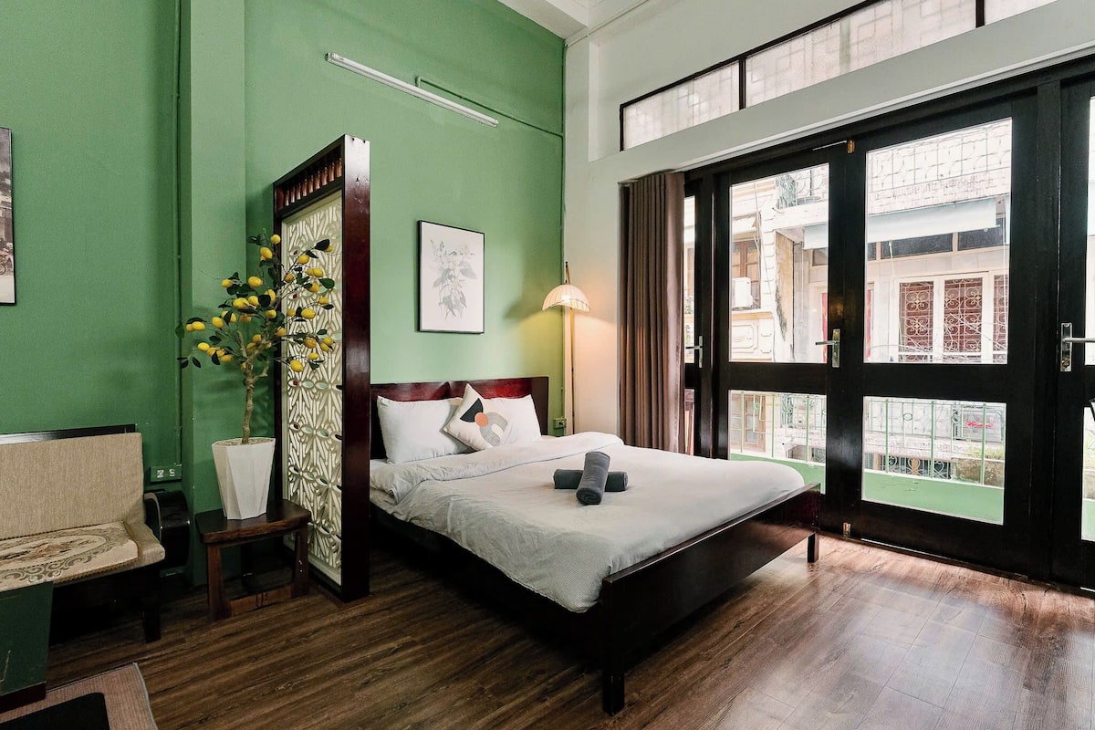 A hotel room with a bed, a sofa, a small table, a lamp, and a window with a view of the street.