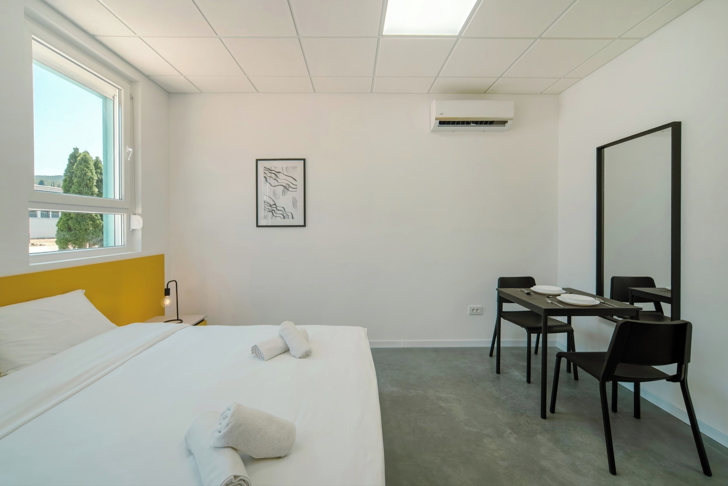 A hotel room with a bed, a desk with chairs, a large mirror, an air conditioner, and a window with a view of trees.