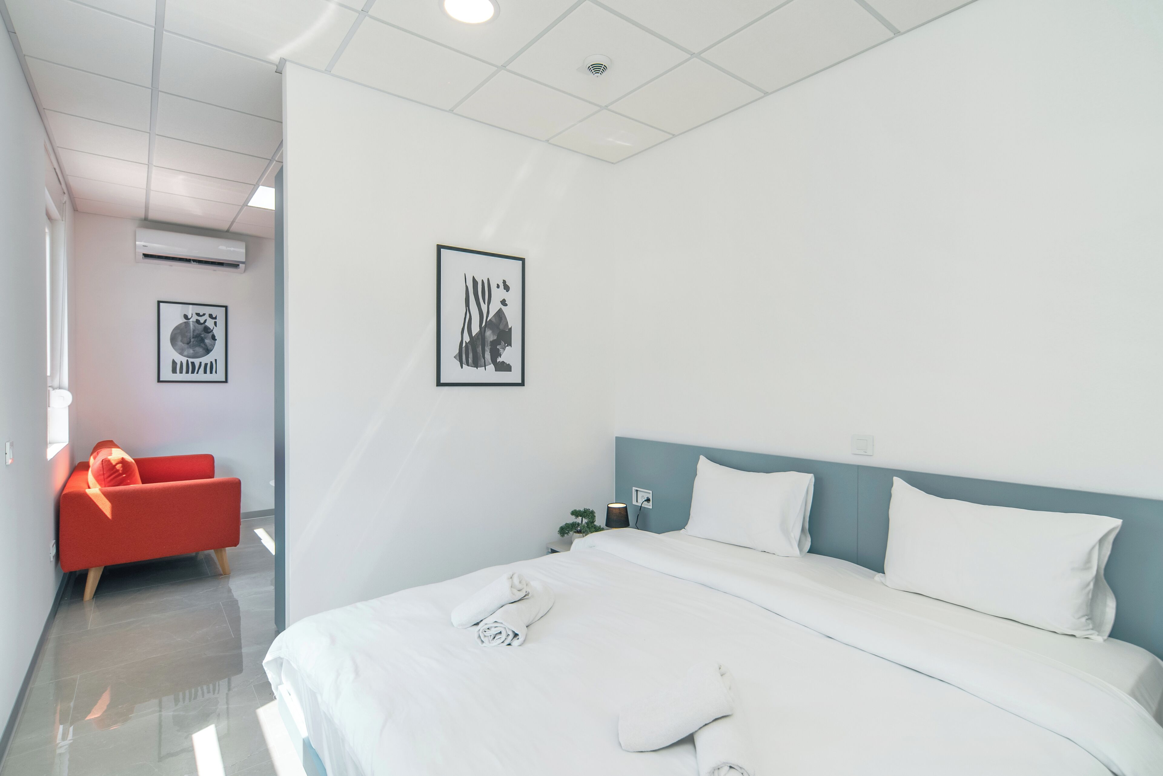 A modern hotel room with a large bed, a red armchair, and two wall-mounted air conditioning units.