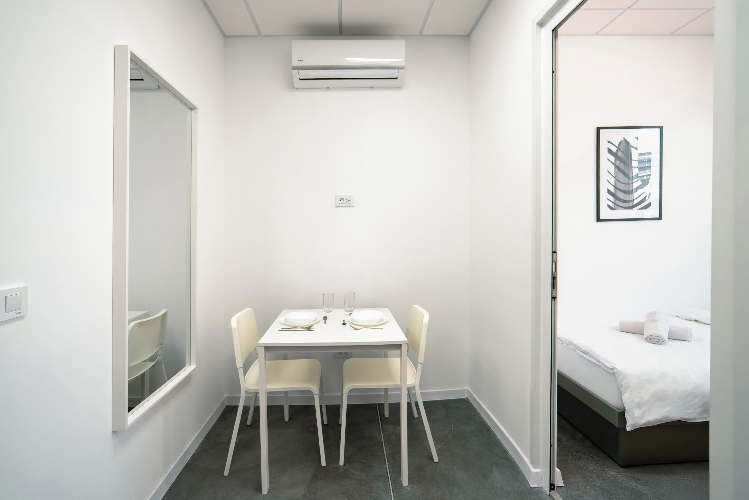 Una habitación pequeña y limpia con una mesa y sillas blancas, cama con ropa de cama blanca, y un cuadro en la pared.