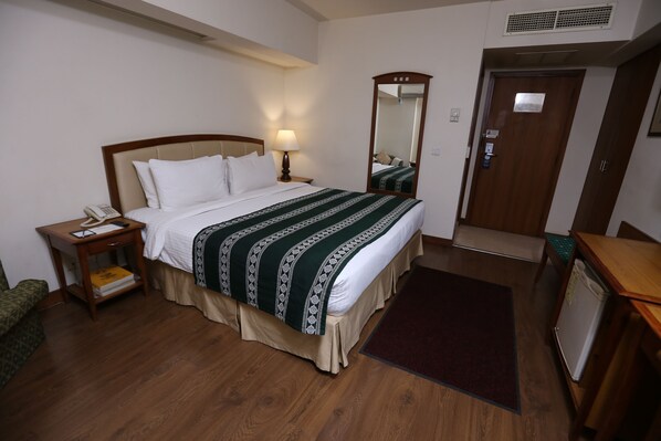 A hotel room with a bed, bedside table, lamp, mirror, door, and a small desk with a phone. - Embassy Inn (Karachi)