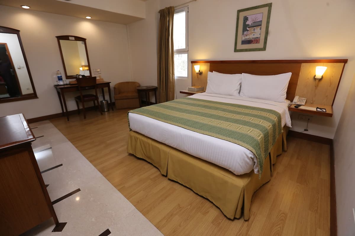 A hotel room with a large bed, a desk with a chair, a small table, a trash can, and a nightstand with a phone and lamp.