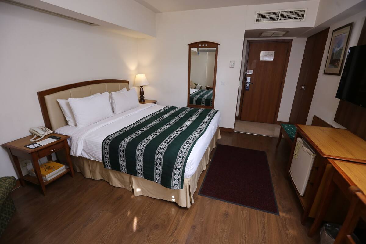 A hotel room with a bed, bedside table with a lamp, a mirror, a door, a desk, and a small refrigerator.