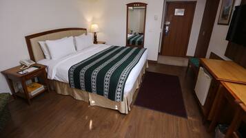 A hotel room with a bed, bedside table with a lamp, a mirror, a door, a desk, and a small refrigerator.