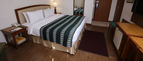 A hotel room with a bed, bedside table with a lamp, a mirror, a door, a desk, and a small refrigerator.