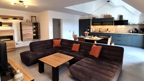 Un salon moderne avec un canapé d’angle, une table basse en bois et un coin repas, la cuisine étant visible au fond.