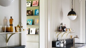 A modern kitchen with white cabinetry, black countertops, and gold accents. There is a built-in bookshelf with books and decorative items, and a small island with a sink and bar tools.