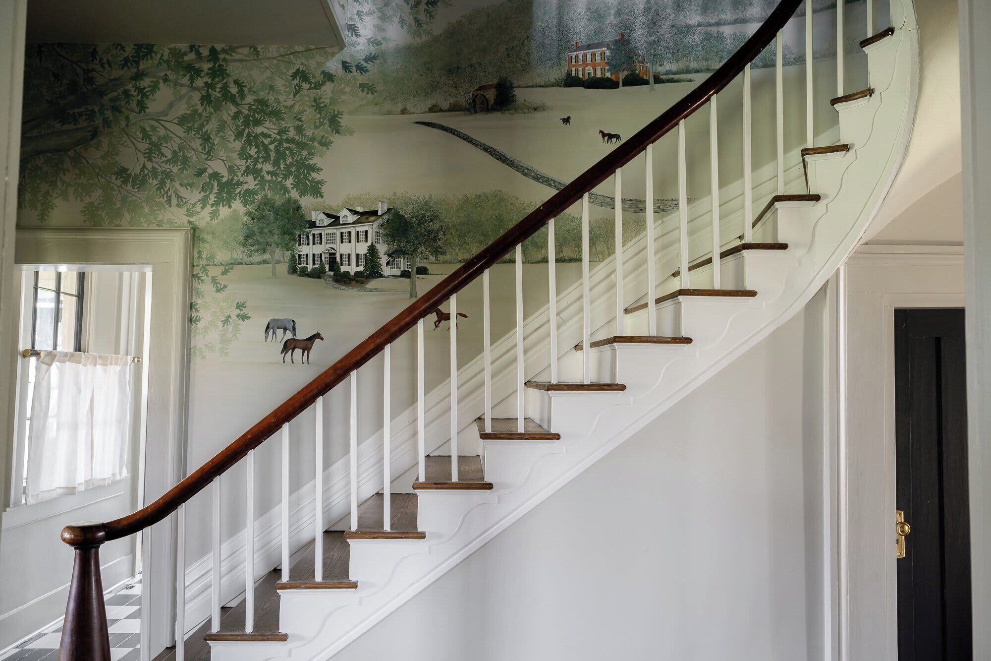 A staircase with white balusters and wooden handrails, leading to a room with a mural of a landscape and a house.