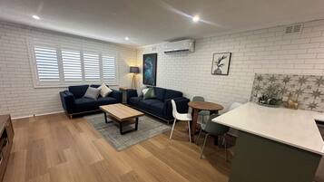 A modern living room with a dark blue sofa, a wooden coffee table, and a small dining area with a white table and chairs.