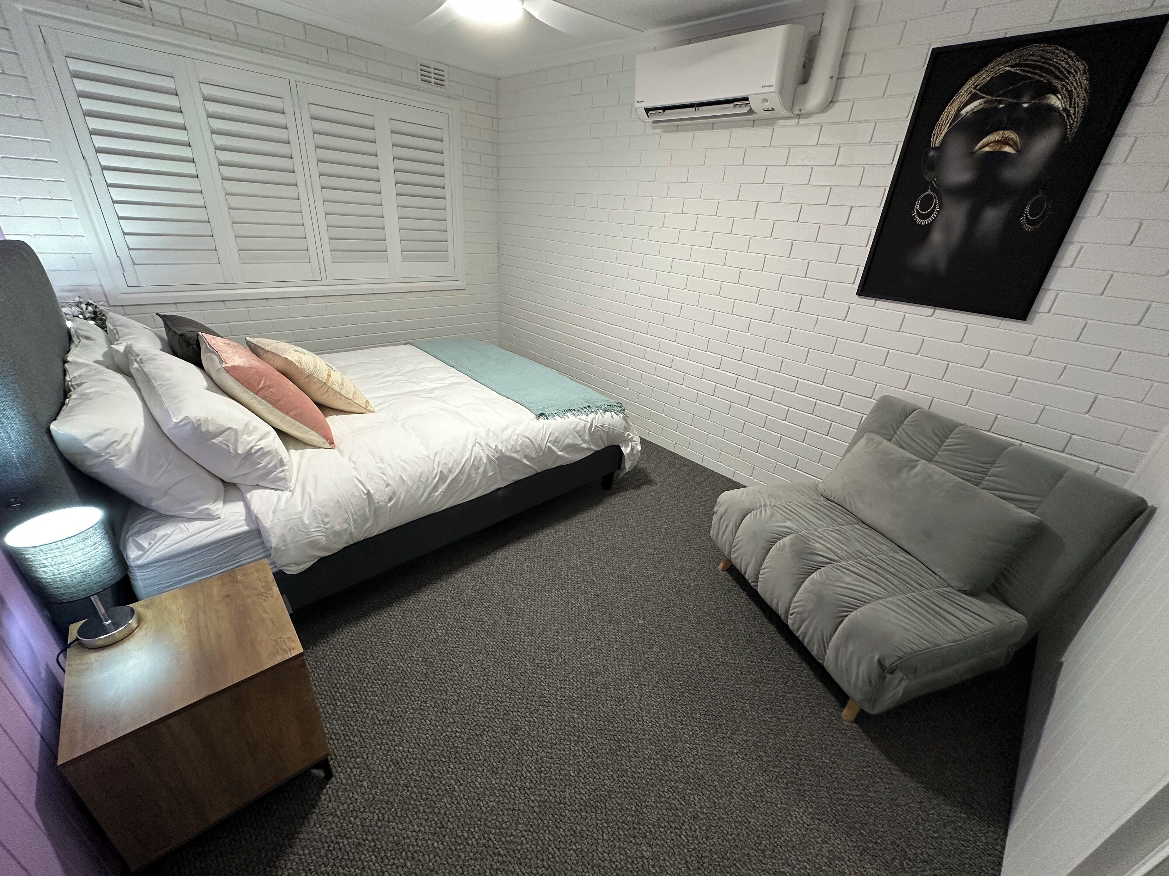 A bedroom with a bed, a grey sofa, a wooden nightstand, and an air conditioning unit on the wall.