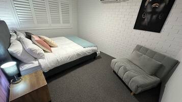 A bedroom with a bed, a grey sofa, a wooden nightstand, and an air conditioning unit on the wall.