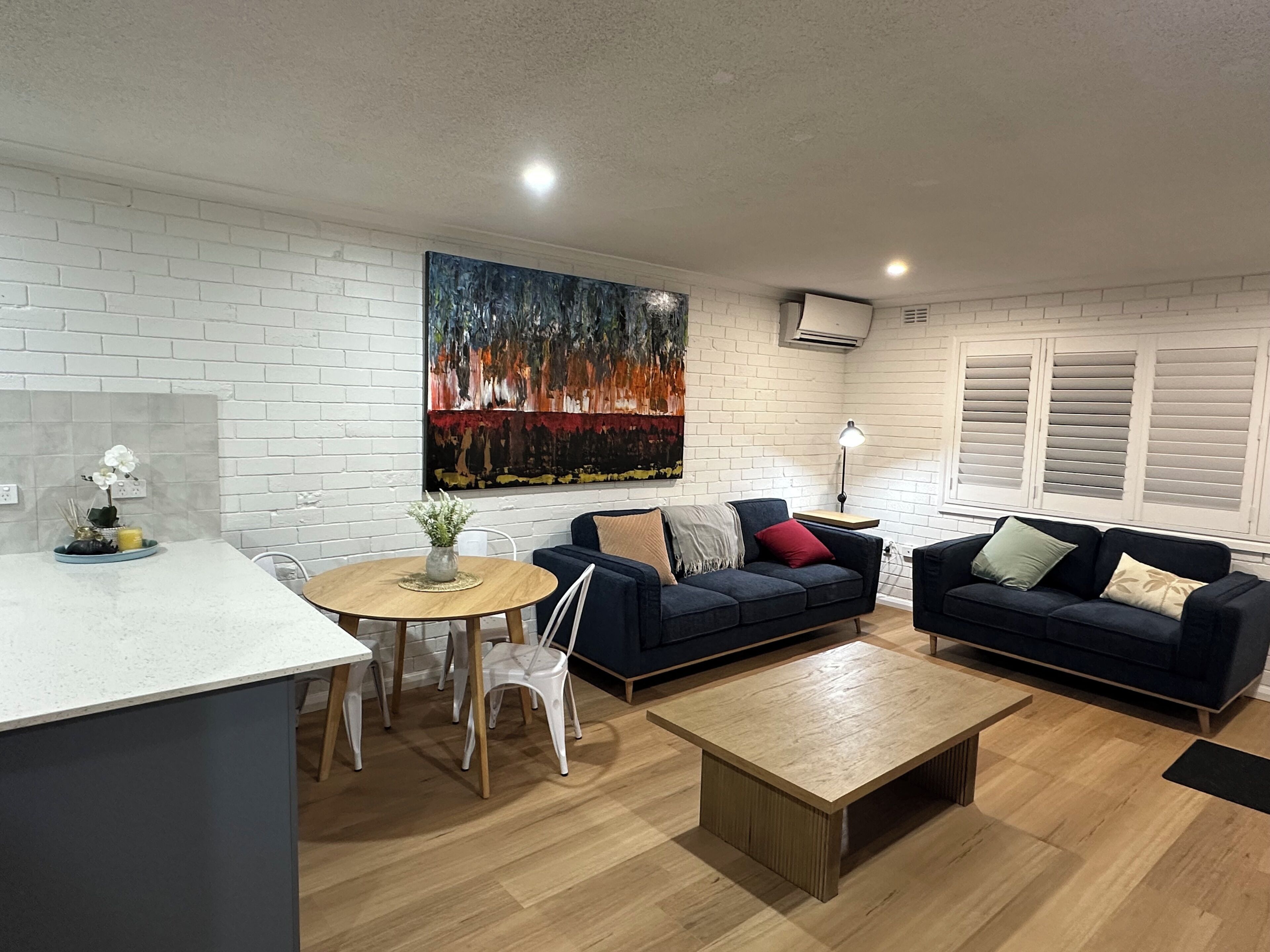 Een moderne woonkamer met een witte bakstenen muur, een groot abstract schilderij, twee donkere banken, een houten salontafel en een kleine ronde tafel met stoelen.