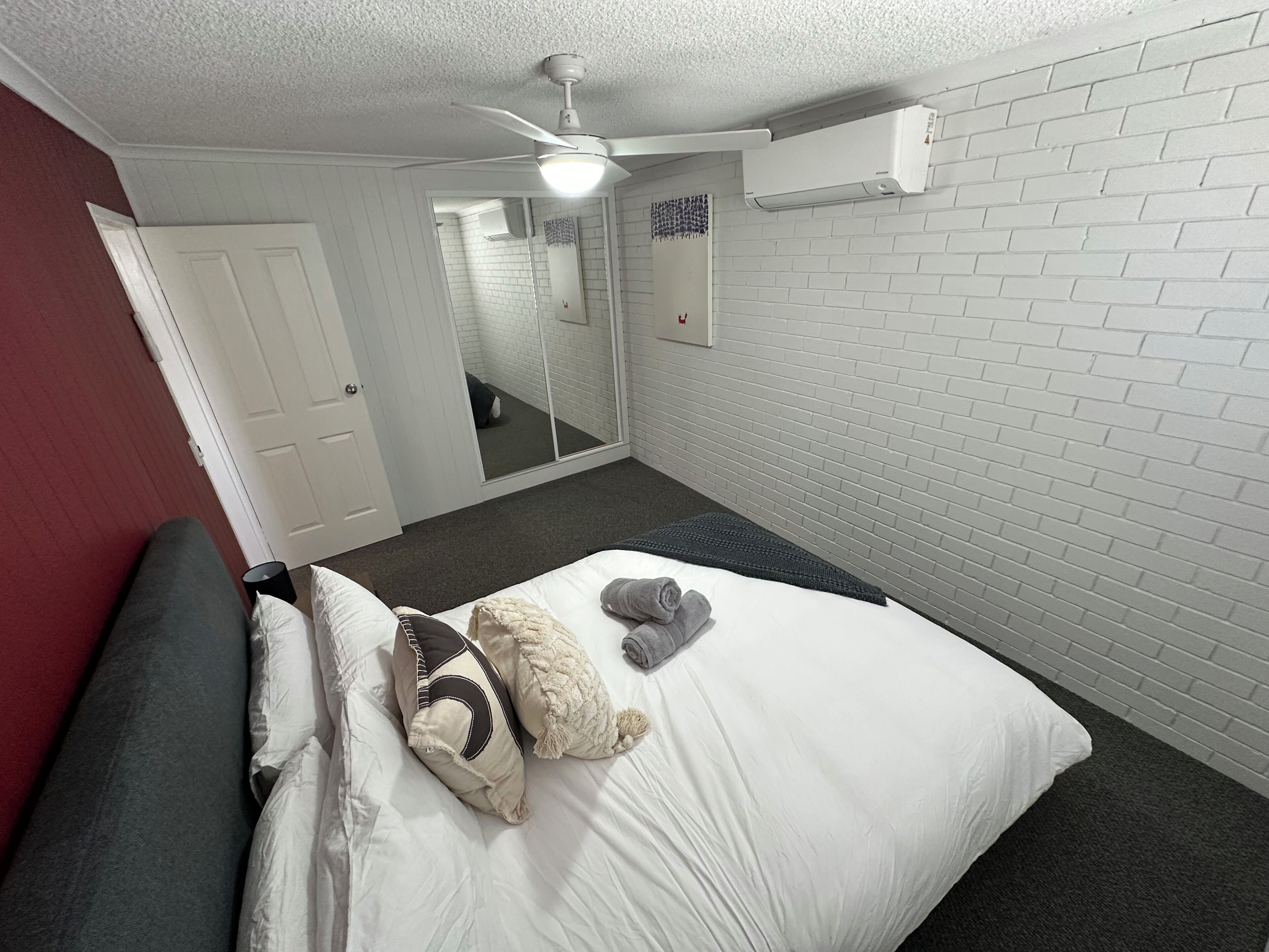 A hotel room with a bed, a ceiling fan, an air conditioning unit, a mirror, and a red wall.