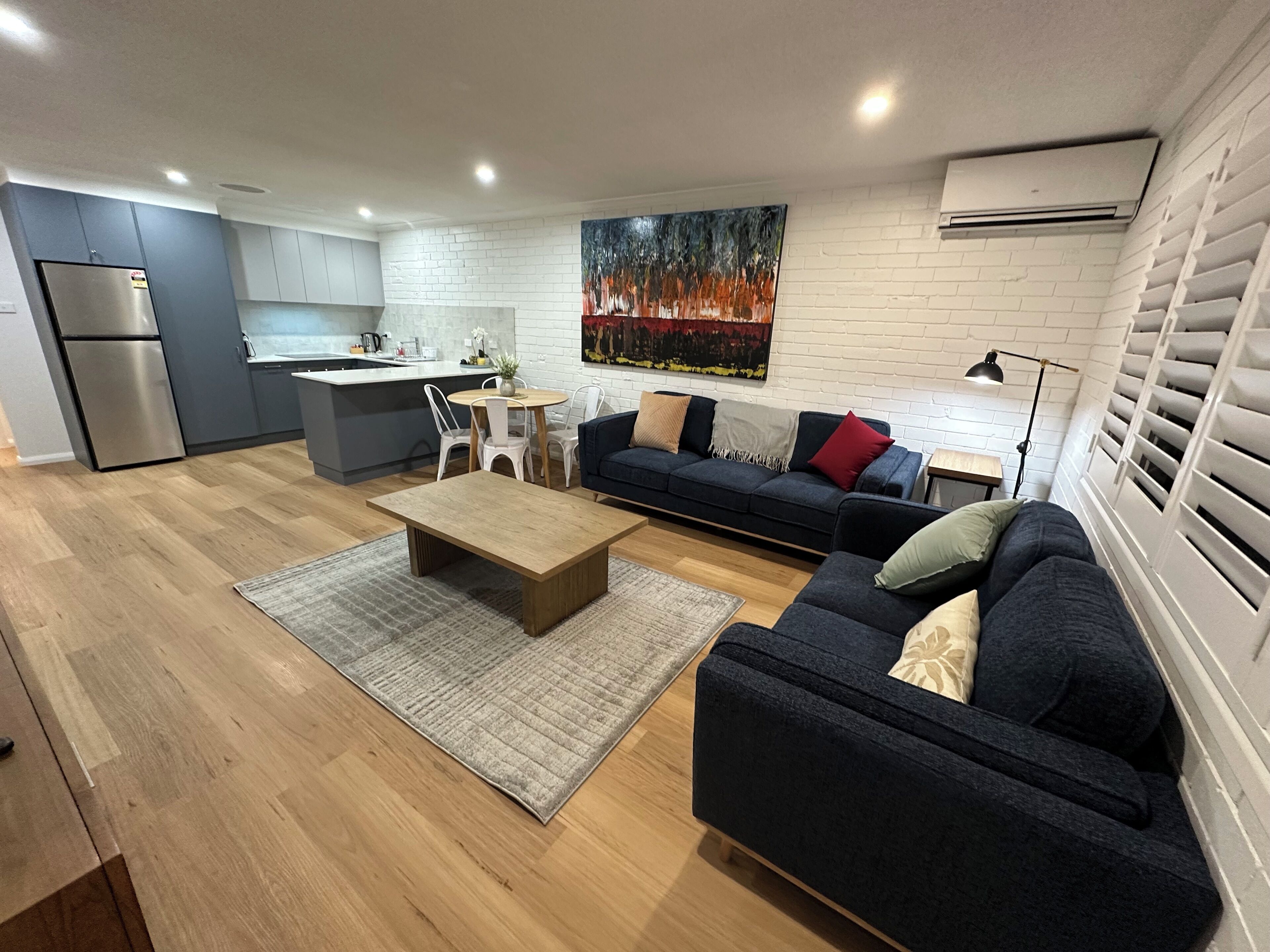A modern living room with a dark blue sofa, a wooden coffee table, and a kitchen area with white cabinets and a stainless steel refrigerator.