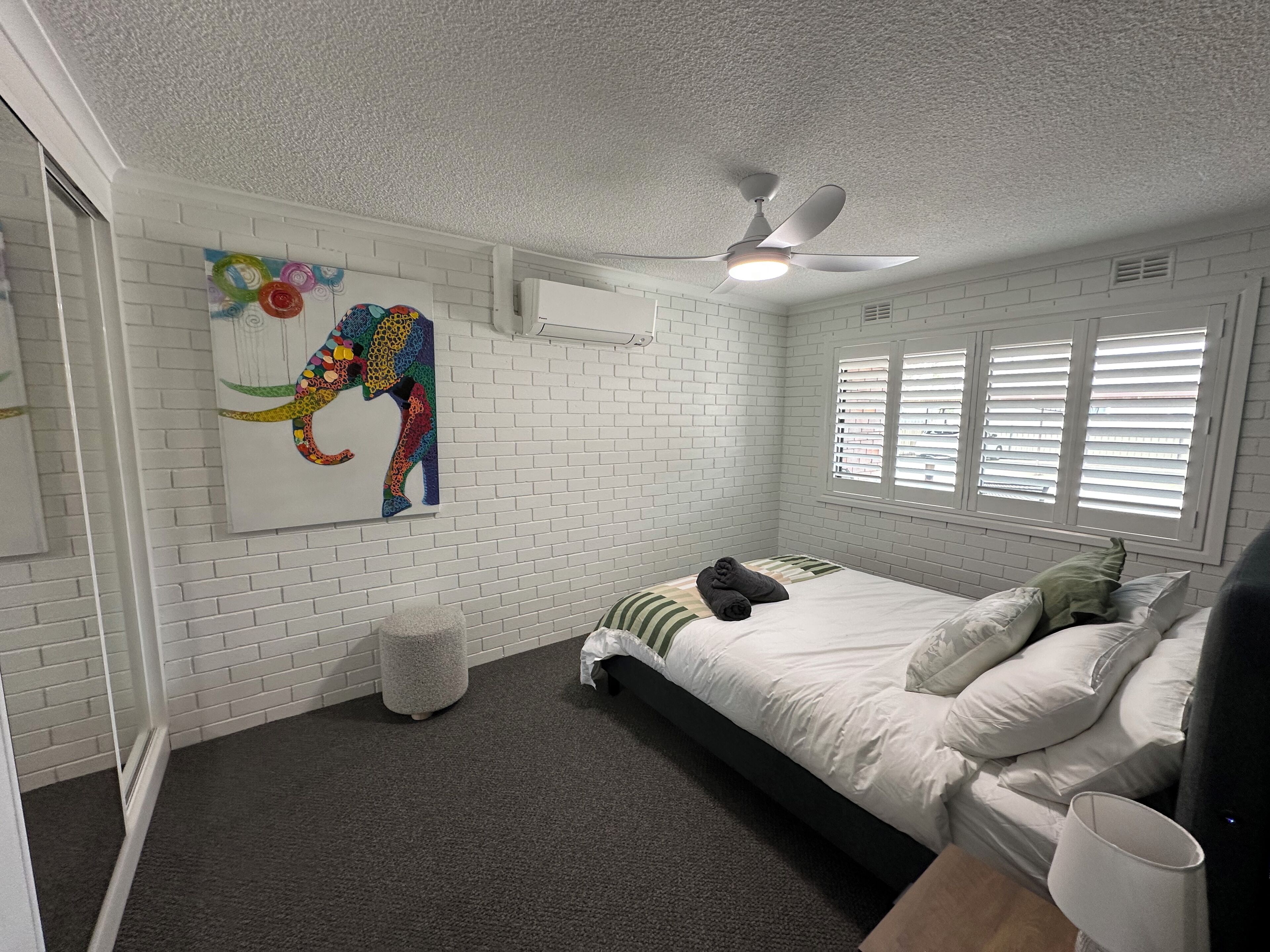Une chambre avec des murs en briques blanches, un ventilateur de plafond, une fenêtre avec des stores, un lit avec une literie et des oreillers blancs, et une œuvre d’art colorée représentant un éléphant accrochée au mur.
