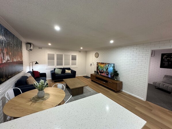 A modern living room with a white brick wall, a black sofa, a wooden coffee table, and a TV on a wooden stand. - Central Cullen Apartments (Wagga Wagga)
