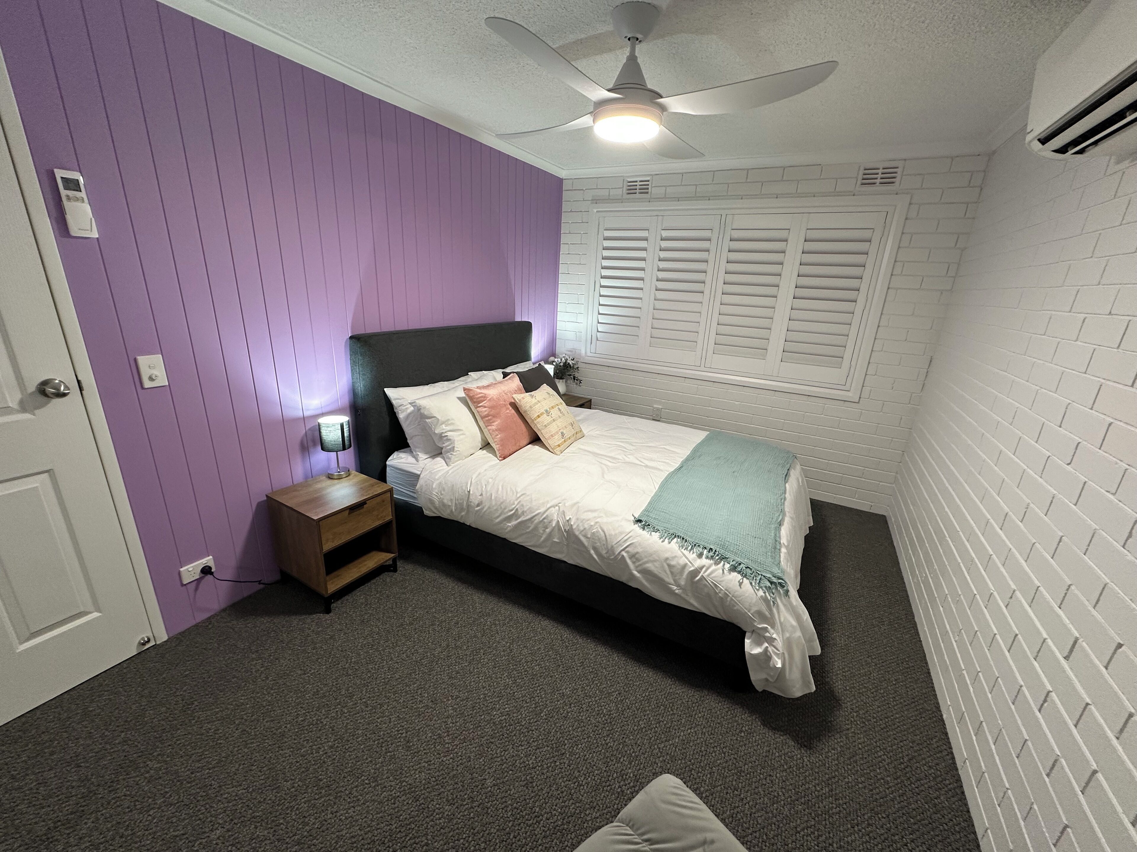 Ein Schlafzimmer mit Bett, Nachttisch, Deckenventilator und Fenster mit Rollläden.