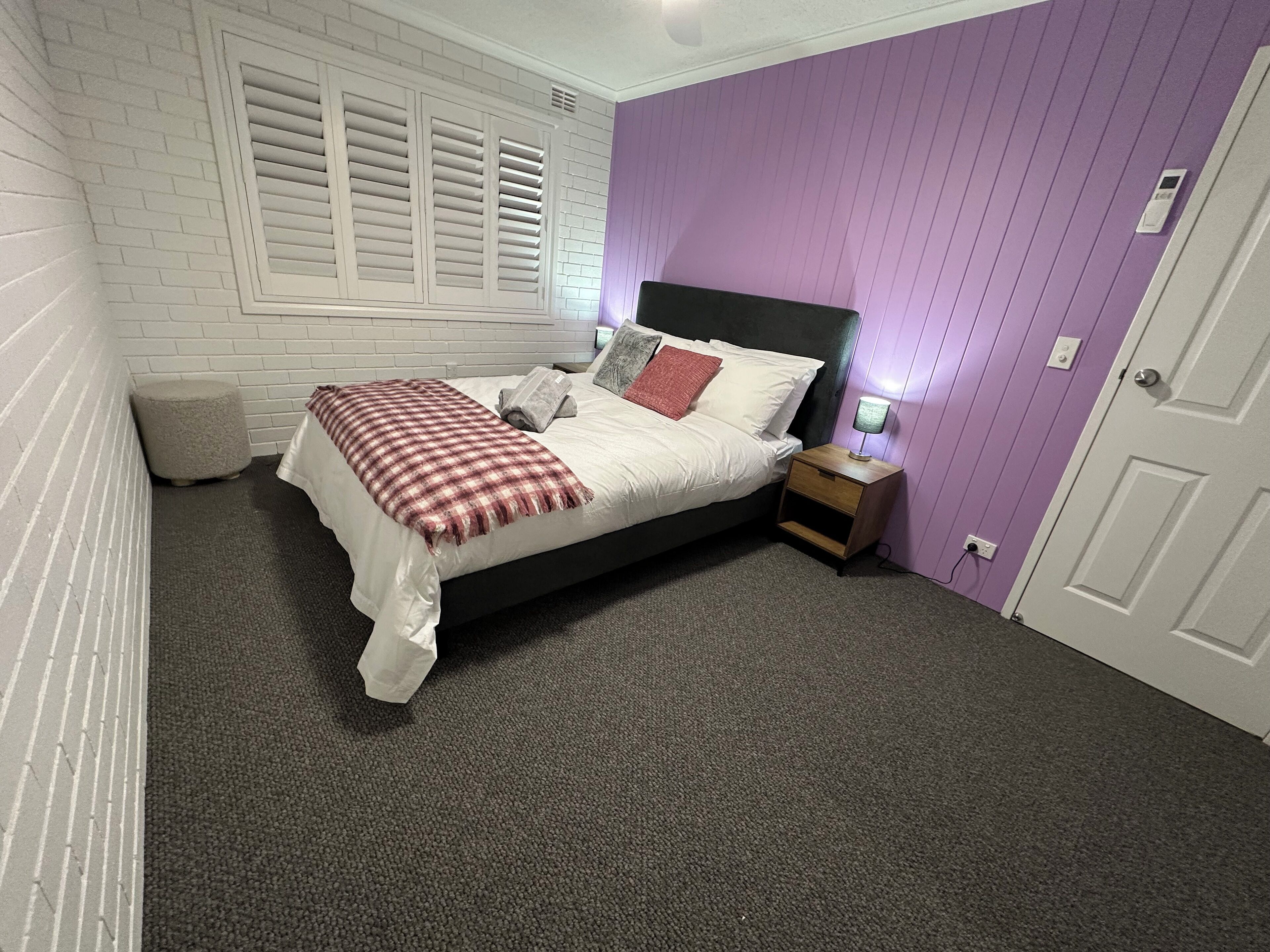 Quarto com cama, mesa de cabeceira, abajur e janela com persianas.